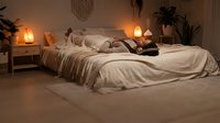 Woman with dark hair and medium skin tone resting on unmade bed with cream linens in warm-lit bedroom with plants and lanterns for relaxation and lifestyle content.