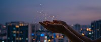 Close-up of an open hand releasing glowing particles against a twilight cityscape with illuminated buildings and bokeh lights in the background.