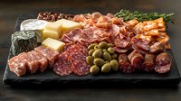 Appetizing charcuterie board with variety of cured meats, cheese, olives and herbs served on black slate plate against dark wooden background. Gourmet food platter.