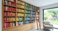 Modern home library with floor-to-ceiling wooden bookshelf filled with colorful books, rolling ladder, and comfortable reading chair by garden window.
