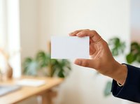 Professional holding blank white card in modern office workspace with natural light and green plants in background, suitable for business branding and corporate identity mockups.