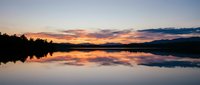 Serene lake at sunset with vibrant orange and purple sky reflecting on calm water surface, surrounded by dark silhouetted trees and mountains in distance.