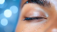 Close up macro shot of closed eye with glitter makeup and sparkles on Asian woman face against blue bokeh background. Professional cosmetics and beauty concept.
