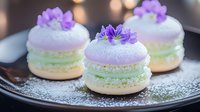 Delicate French macarons in pastel mint green and lavender topped with purple edible flowers, served on dark plate with powdered sugar dusting.