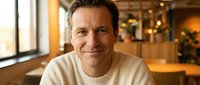 Middle-aged Caucasian man in cream sweater smiling warmly at camera in cozy cafe setting with soft lighting and blurred background for lifestyle content.