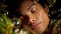 Young African American woman with closed eyes and serene expression surrounded by glowing bubbles and green foliage in dreamy natural lighting for wellness and beauty concepts.