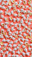 Fresh white popcorn kernels scattered across vibrant coral red background for movie night entertainment and snack food concepts.