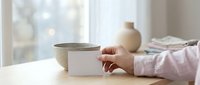 Woman's hand holding white ceramic bowl on wooden table near window with soft natural light creating peaceful morning atmosphere for mindful eating.
