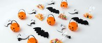 Halloween party decorations with orange pumpkin buckets, black bats, white ghosts and colorful candy scattered on white background for festive celebration.