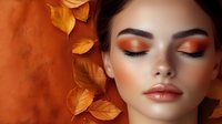 Young woman with autumn makeup and closed eyes surrounded by golden fall leaves on orange background, demonstrating professional cosmetics application.