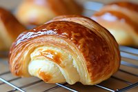 Fresh baked golden croissant with flaky layered texture and glossy butter glaze cooling on metal rack, extreme close up view showing delicate pastry details.