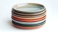 Stack of handcrafted ceramic plates in pastel colors featuring coral pink, sage green, and neutral tones against white background, showcasing artisanal pottery design.