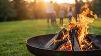 Crackling campfire flames in metal fire bowl with blurred family gathering in sunny park background during golden hour outdoor recreation.