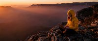 Hiker in yellow jacket sits on mountain cliff edge at golden sunrise, overlooking misty valley landscape below.