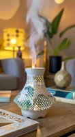 Decorative white ceramic aroma diffuser with intricate lattice pattern emitting steam, creating cozy atmosphere with warm lighting and houseplants in background.