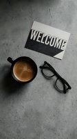 Welcome card, black coffee mug, and eyeglasses on gray concrete surface, flat lay composition for business or lifestyle use.