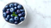 Fresh blueberries in white bowl on marble surface with mint leaf garnish for healthy eating and organic food concepts.