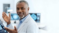 African American male doctor in white coat gesturing while explaining medical procedure in modern hospital with diagnostic equipment background.
