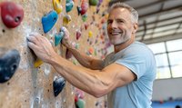 Mature athletic man with gray hair and beard smiling while climbing on indoor rock wall with colorful holds at climbing gym, demonstrating active lifestyle and fitness.