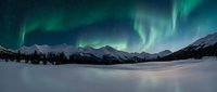Spectacular aurora borealis dancing over snow-covered mountain landscape with frozen lake creating magical winter night scene for nature photography and travel content.