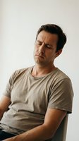 Middle-aged Caucasian man experiencing stomach pain or nausea, holding abdomen with concerned expression, wearing casual beige t-shirt against neutral background.