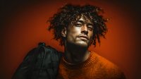 Young African American man with curly hair wearing orange sweater carrying backpack against warm gradient background for lifestyle and education concepts.