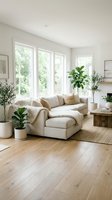 Bright modern living room with cream upholstered sectional sofa, natural wood flooring, large windows with greenery views, potted plants, and minimalist decor for home interior design inspiration.