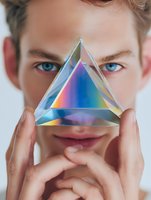 Young man with blue eyes holding prismatic crystal triangle creating rainbow light effect against white background for spiritual or wellness concept.