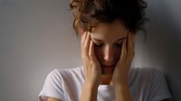 Young woman in white t-shirt experiencing headache or emotional distress, holding head in hands against neutral background, depicting mental health concept.
