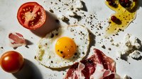 Fresh breakfast ingredients scattered on white surface with sunny side up egg, prosciutto slices, tomatoes and black pepper, captured from above in natural light.
