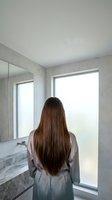 Woman with long brown hair in silk robe standing by modern bathroom window looking outside in morning light for wellness and self-care concepts.