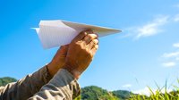 Hand holding white paper airplane against bright blue sky with clouds, symbolizing freedom, dreams, and new beginnings in outdoor summer setting.