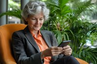 Senior businesswoman in gray suit and coral blouse sitting in orange armchair, smiling while using smartphone, with lush green plants in background.