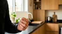 Person holding blank white mug in modern kitchen with wooden cabinets and natural light, suitable for mug mockups, branding, and beverage marketing campaigns.