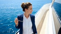 Woman in business casual attire standing on luxury sailboat deck overlooking blue ocean waters, holding anchor chain, representing maritime travel, yacht lifestyle, and professional leisure activities.