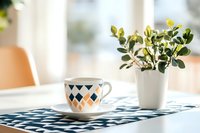 Morning coffee cup with geometric pattern on blue tablecloth next to white vase with fresh yellow flowers in sunlit modern minimalist kitchen interior.
