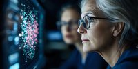 Senior Caucasian woman data scientist wearing glasses analyzes digital visualization on screen while collaborating with colleague in dark office environment.