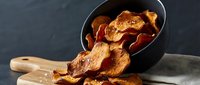 Crispy golden sweet potato chips spilling from black bowl onto wooden cutting board, healthy snack alternative for cooking and food photography.