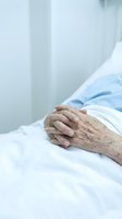 Elderly patient hands folded on hospital bed with white sheets in medical care setting for healthcare and treatment concepts.