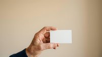 Close-up of mature male hand holding blank white business card against neutral beige background for corporate branding and professional identity mockup purposes.