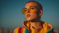 Young woman with short blonde hair wearing oversized round yellow glasses and colorful hoodie against sunset sky. Golden hour portrait with warm lighting.