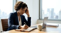 Stressed African American businesswoman holding head while working at office desk with laptop and documents in modern corporate environment.