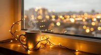 Cozy evening scene with steaming coffee mug and warm fairy lights on windowsill overlooking blurred city lights creating peaceful atmosphere for relaxation.
