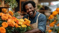 Smiling African American male florist arranging vibrant orange chrysanthemums in flower shop, wearing apron while working with fresh autumn blooms for seasonal arrangements.