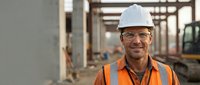 Smiling mature Caucasian construction worker in white hard hat and orange safety vest at building site with concrete structure and machinery in background.