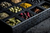Assorted aromatic spices and herbs in black compartment box, including cardamom, turmeric, star anise, peppercorns, and bay leaves on dark background.