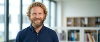 Smiling middle-aged Caucasian businessman with curly hair and beard in navy shirt standing in modern office with bookshelves for corporate headshot.