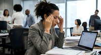 Stressed businesswoman with headache at office desk while colleagues work in background. Workplace pressure and mental health concept for corporate wellness.