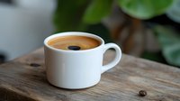 Fresh espresso with crema in white ceramic cup on rustic wooden table with green plant leaves in soft natural lighting, perfect for cafe menu or blog.