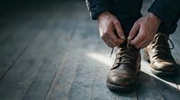 Man tying brown leather work boots on wooden floor preparing for outdoor activity or labor job with dramatic lighting and weathered hands.